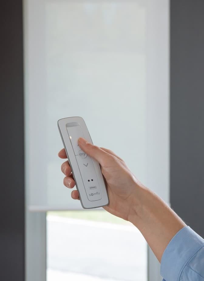 A hand pressing the lower button on a Somfy remote, the blind descending behind it